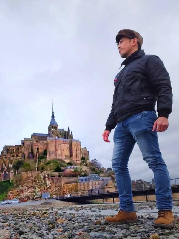 Traveler standing on a rocky shore with Mont Saint Michel rising behind him under an overcast sky.