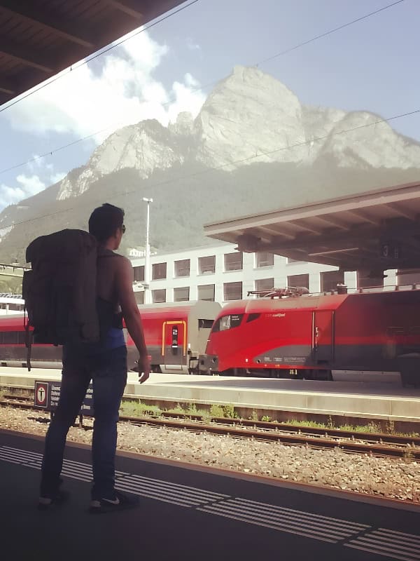 Traveler with a backpack waiting beside a red train at a European mountain railway station with snow covered peaks behind him.