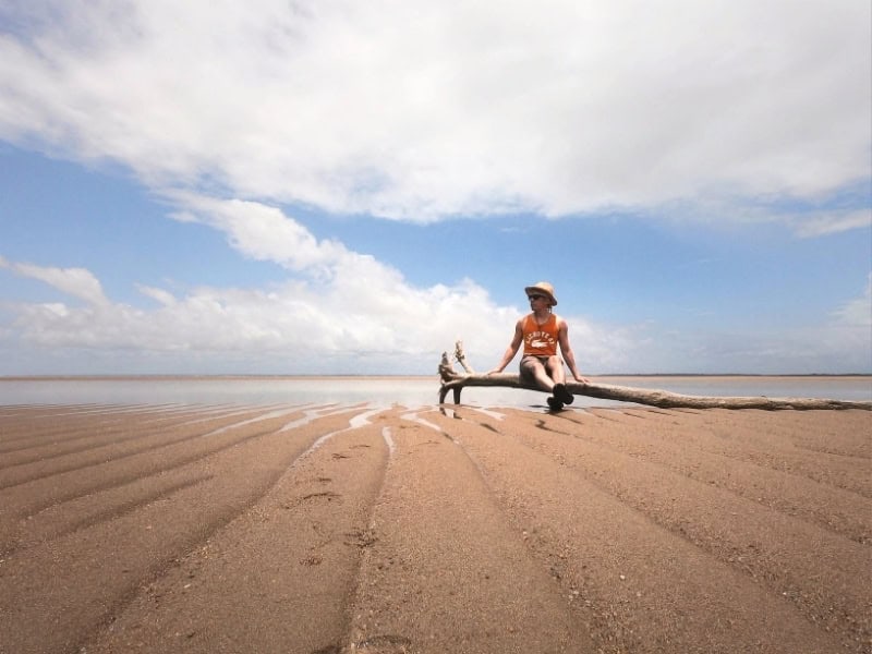 Traveler sitting on driftwood across a wide sandy beach at low tide beneath a bright blue sky and broad white clouds.