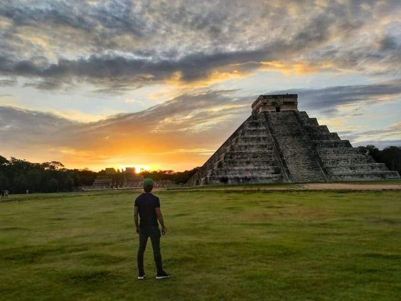 Traveler standing in a grassy field facing Chichen Itza at sunset with dramatic clouds overhead.