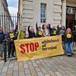 PILS and Stop Whitehead Oil Terminal campaigners stand on the steps of the Royal Courts of Justice, with a large yellow banner, celebrating their legal win!