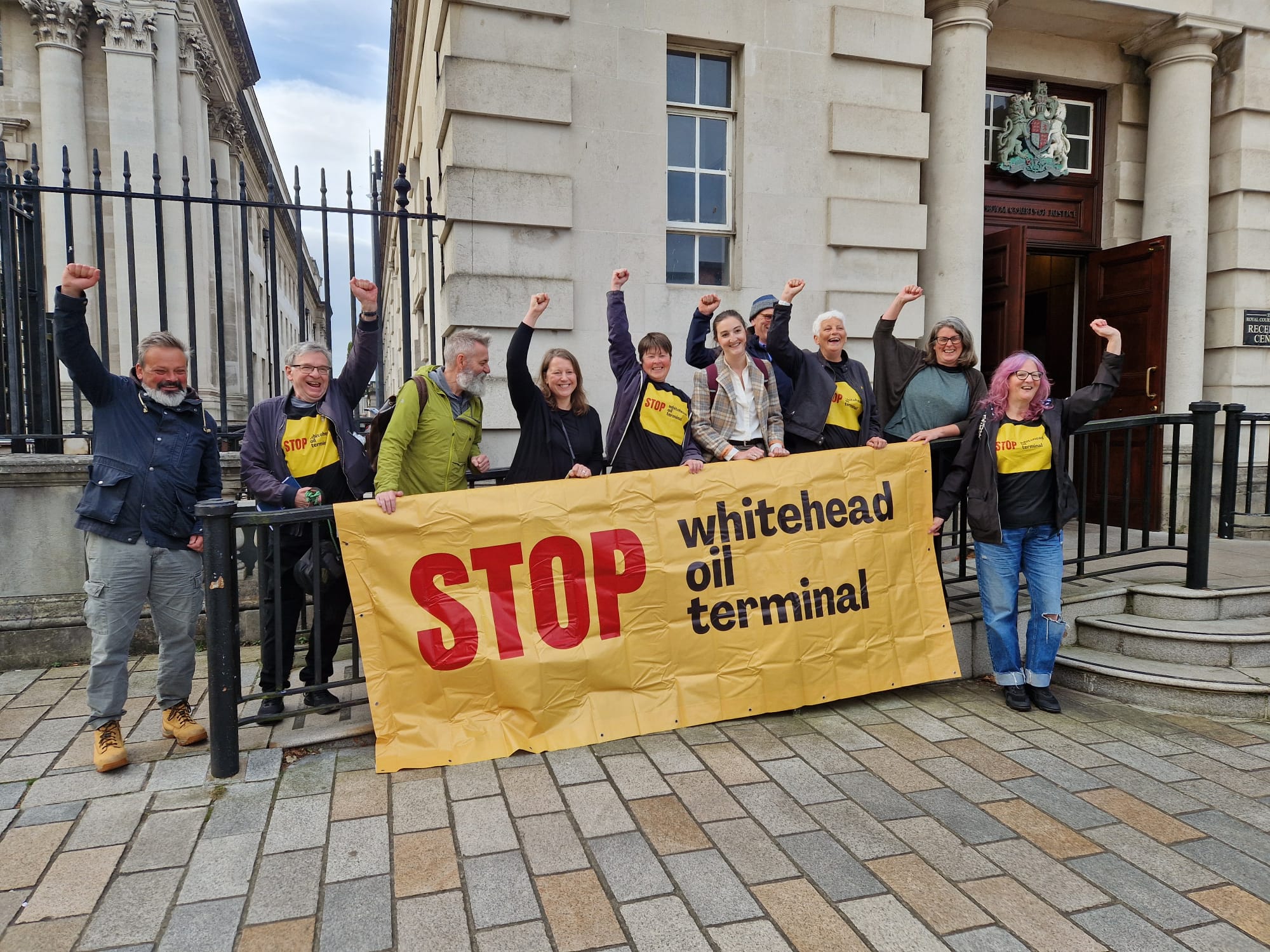 PILS and Stop Whitehead Oil Terminal campaigners stand on the steps of the Royal Courts of Justice, with a large yellow banner, celebrating their legal win!