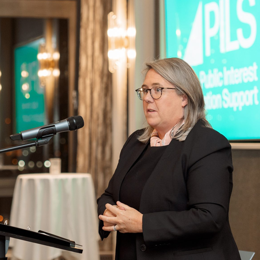 Lady Chief Justice of Northern Ireland, Dame Siobhan Keegan, is standing behind a podium. She is a white woman with shoulder-length ash blonde hair and glasses. She is speaking confidently into a microphone, addressing the audience at PILS' 15h Anniversary event.