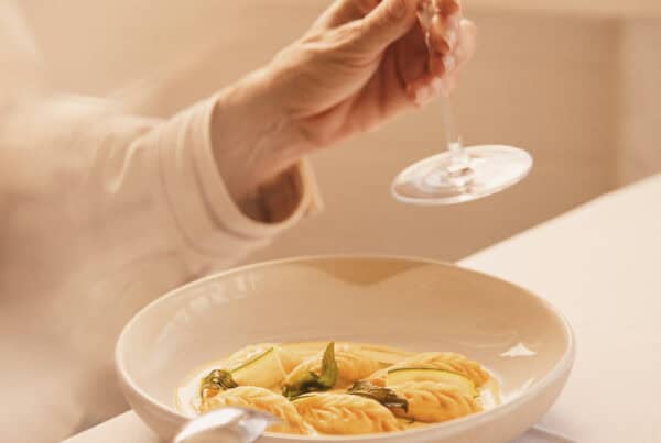 A woman holding a glass of white wine above a plate of pasta on a dining table.