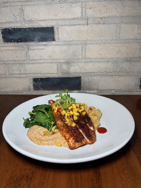 Roasted garlic hummus served with grilled fish, greens, and corn on a white plate.