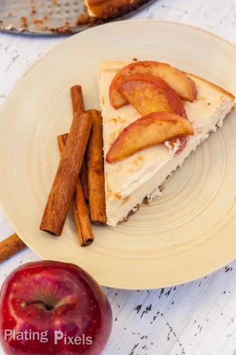 Slice of apple cider cheesecake with apple slices and cinnamon sticks on a plate.