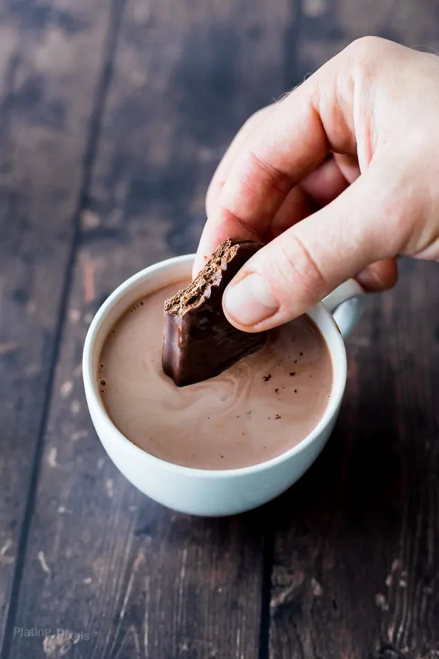 Hand dipping chocolate cookie into hot chocolate for a cozy drink.