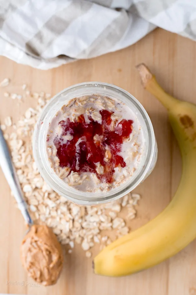 Vegan peanut butter and jelly overnight oats in a glass jar with banana on a wooden surface.