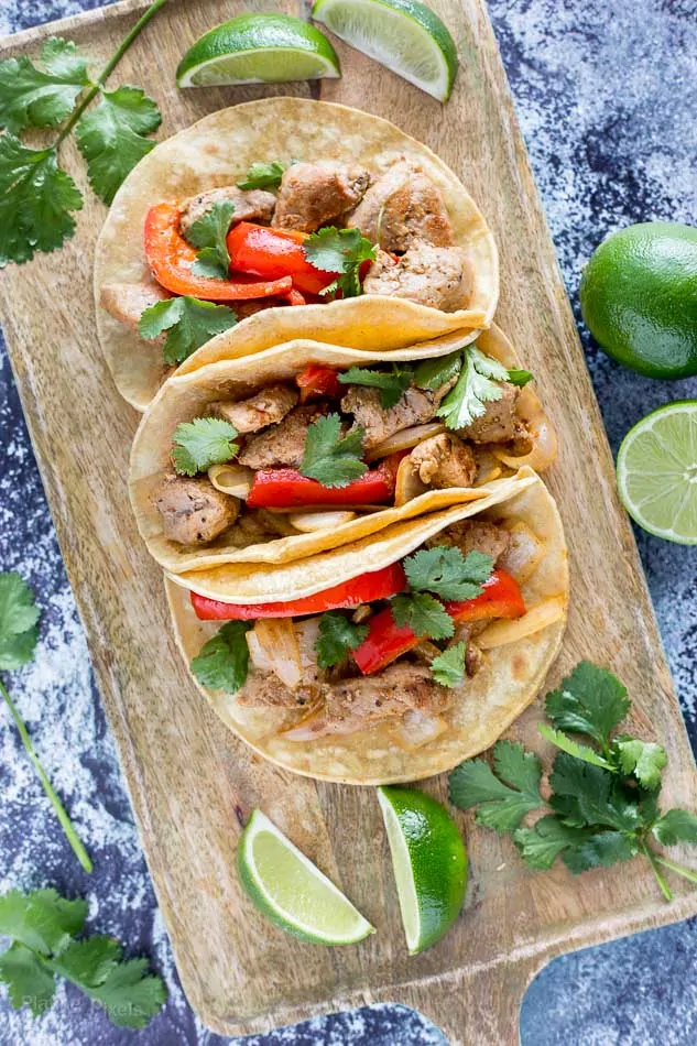 Pork fajita tacos with peppers, cilantro, and lime wedges on a wooden board.