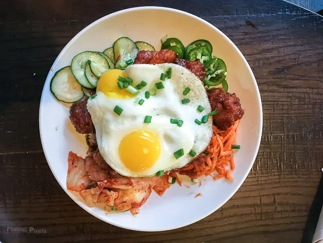 Korean breakfast bowl with fried eggs, cucumbers, and kimchi on a white plate.