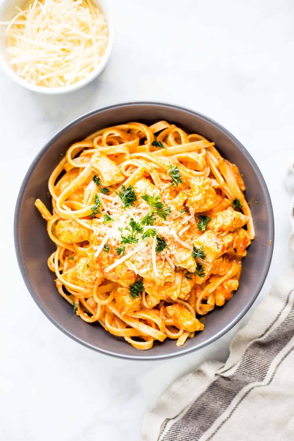 Rosé pasta with chicken and herbs in a black bowl, garnished with cheese and parsley.