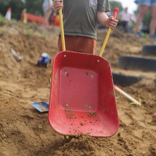 Child playing in a sandpit with a red toy wheelbarrow at Playful Paths outdoor play area.