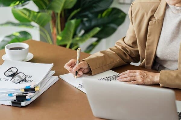 High-quality planner for all PLR materials on a wooden desk with a person writing, surrounded by notebooks, a laptop, and a cup of coffee, ideal for digital marketers and content creators.