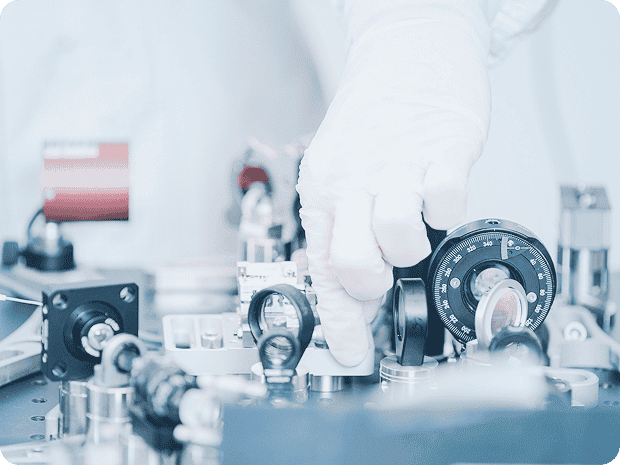 A gloved hand making fine adjustments to a set of precision optical or measurement instruments on a laboratory workbench, surrounded by lenses, dials, and metal components.