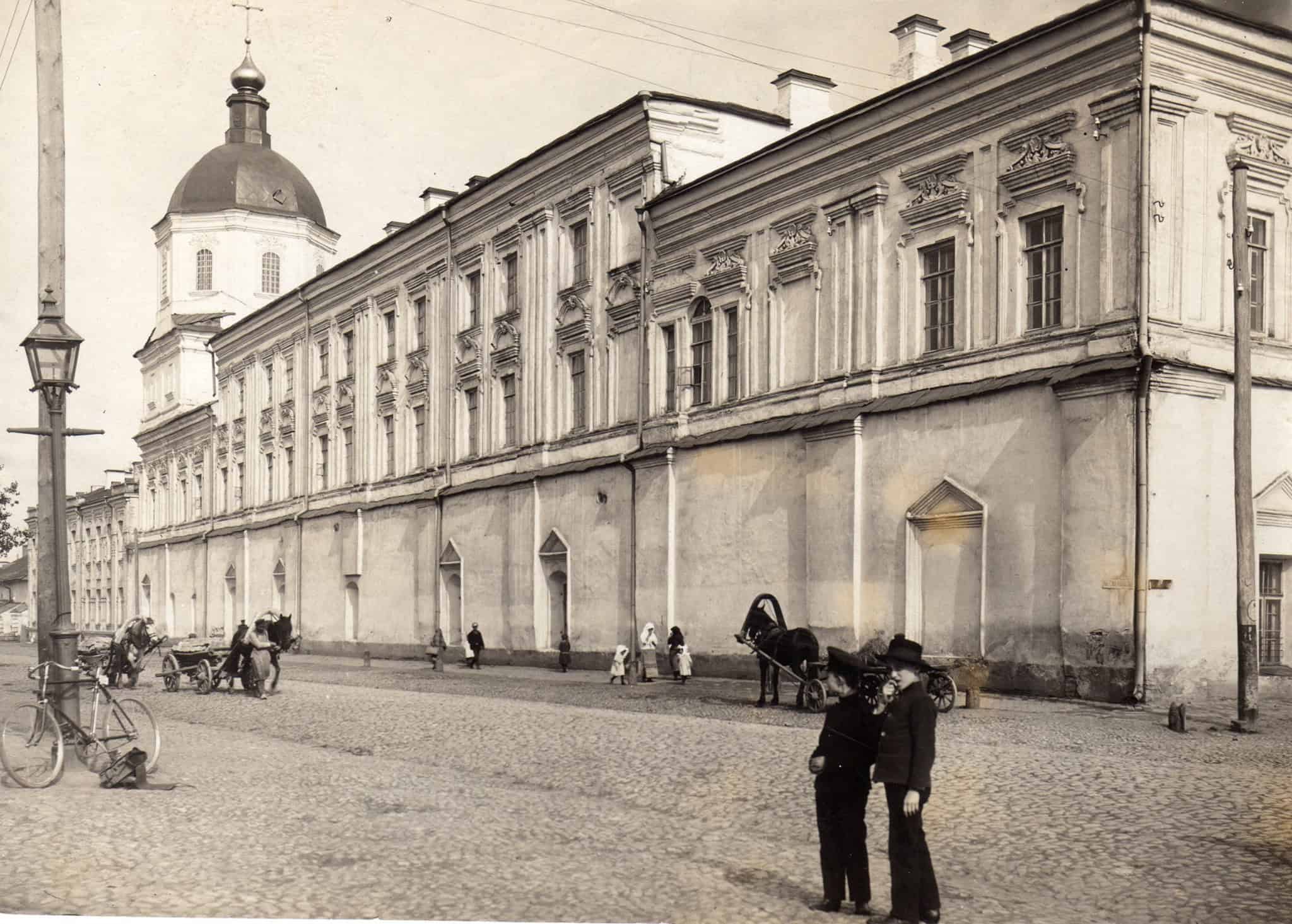 Історична фотографія старовинної будівлі на вулиці з кінськими екіпажами.