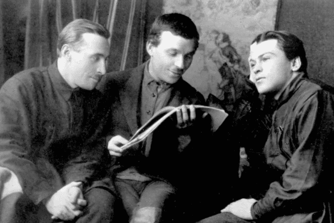 Микола Куліш у компанії двох чоловіків, читає книгу, чорнобіле фото.
