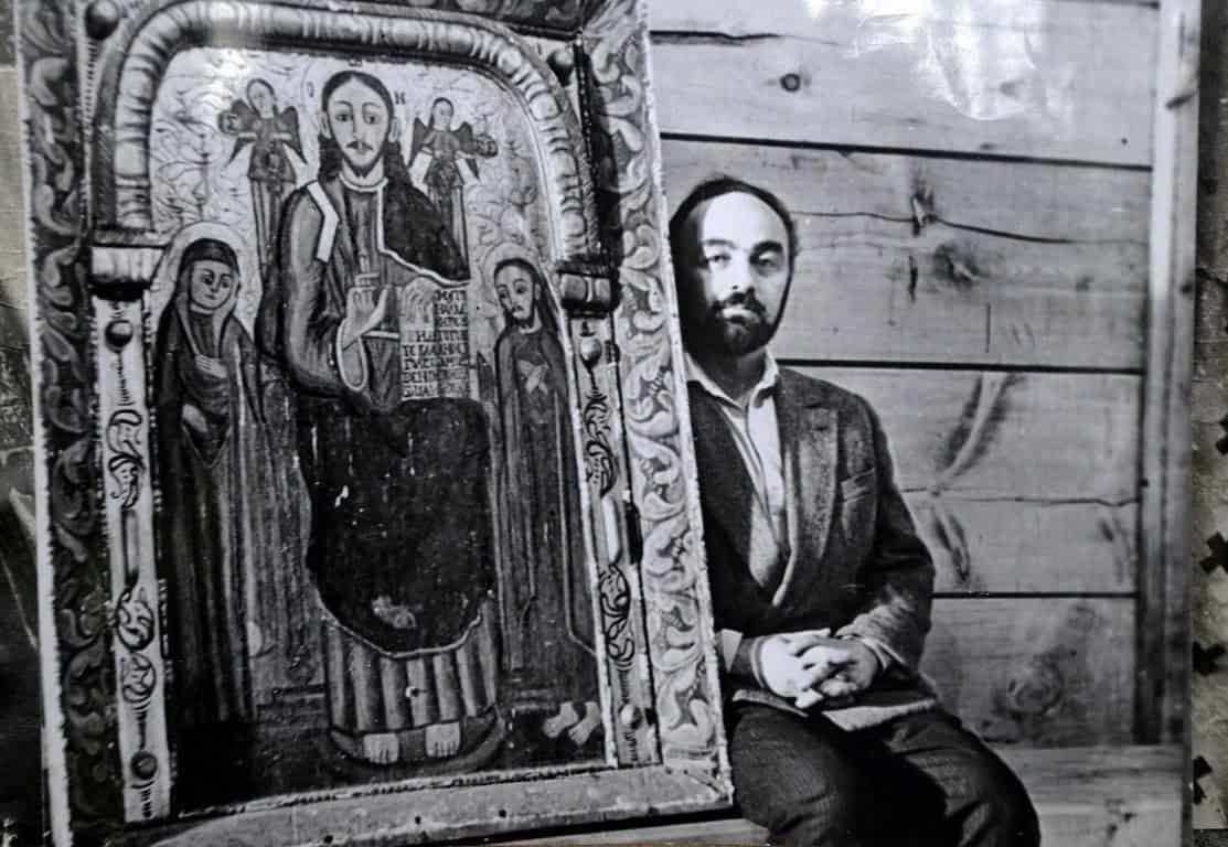 Чоловік поруч із іконою на дерев'яній стіні, історична фотографія.