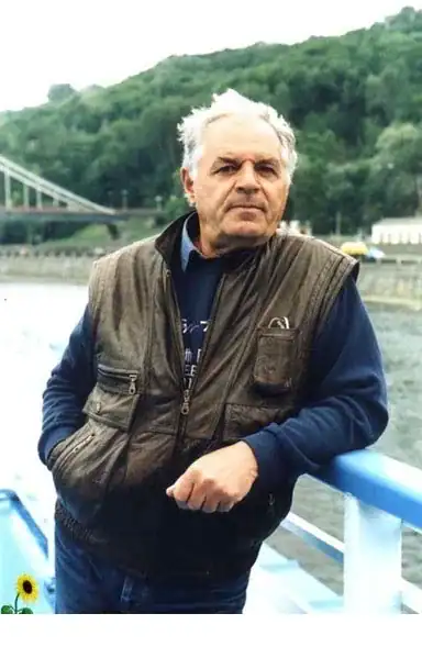 Чоловік на фоні річки та зелених пагорбів, одягнений у жилет і куртку.