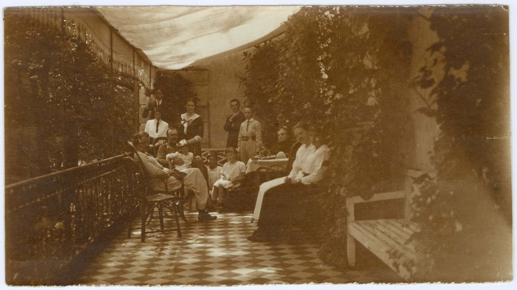 Група людей на відкритій терасі з квітами та деревами, історична фотографія.
