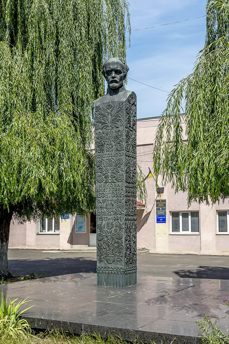 Пам'ятник з бюстом на високій стелі серед зелених дерев у місті.
