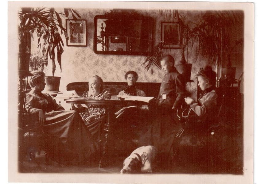 Група людей у кімнаті з рослинами та фотографіями на стінах, історична фотографія.