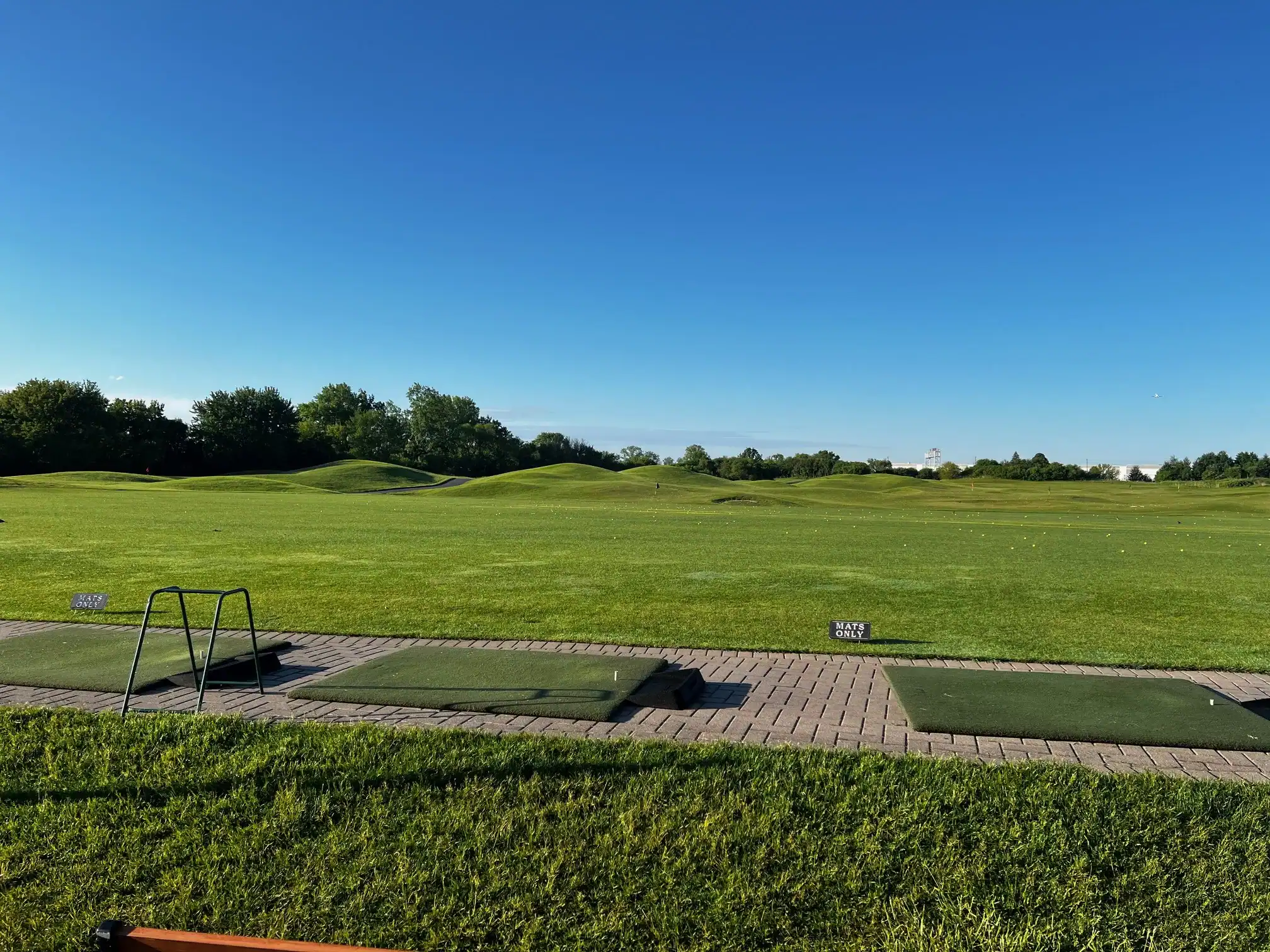 A wide view of a golf driving range with green hitting mats in the foreground, a vast green field with scattered golf balls, and a clear blue sky with a distant airplane.