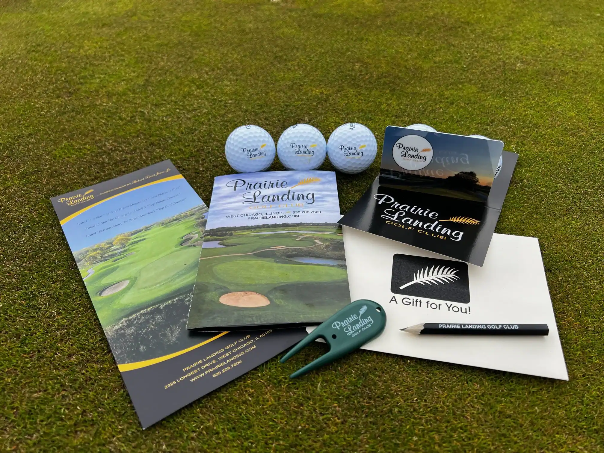 Promotional items from Prairie Landing Golf Club, including three branded golf balls, various brochures with golf course images, a green divot tool, and a black pencil, all displayed on green grass.