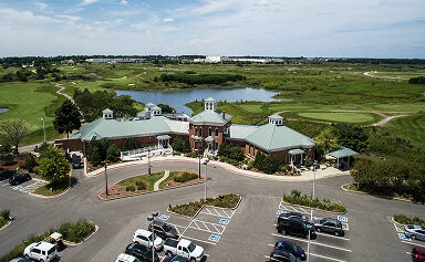 Aerial view of a large brick clubhouse with a green roof, surrounded by a parking lot, golf course fairways, a lake, and marshland under a blue sky.
