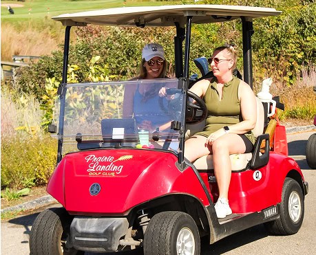 Two smiling women in a red golf cart on a sunny golf course. The driver wears olive green, and the passenger wears a blue top and white cap.