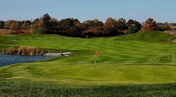 A scenic golf course with a red flag on the green, a pond with reeds, and trees displaying autumn foliage under a clear blue sky.