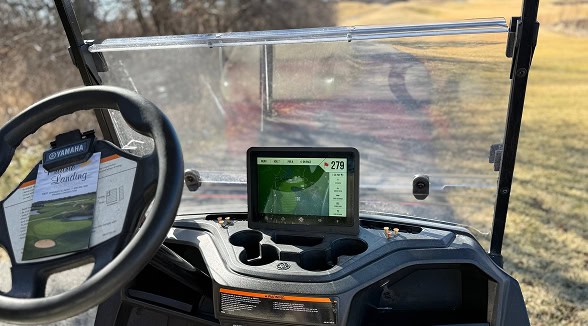 Interior view of a golf cart dashboard with a steering wheel, a 'Sterling Landing' scorecard, and a digital screen displaying a golf course map with a 279-yard distance. A golf course is visible through the windshield.