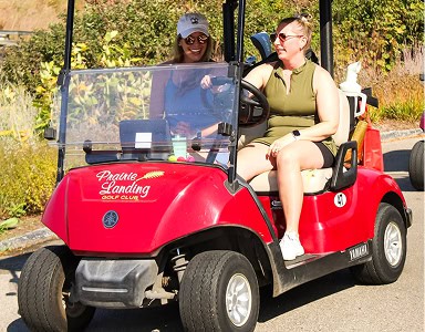 Two women smiling and riding in a red golf cart at Prairie Landing Golf Club on a sunny day. The driver wears a green top, and the passenger wears a blue top and white cap.