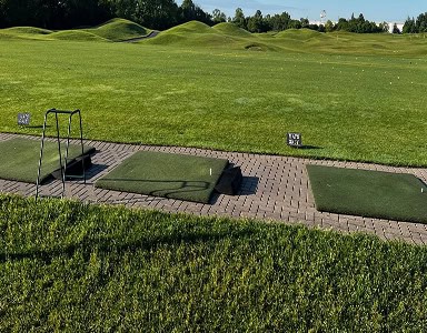 A golf driving range with three artificial turf hitting mats on a paved area. The range features vast green grass, scattered golf balls, and rolling green hills in the background under a clear sky.