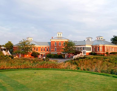 A large red brick clubhouse with white cupolas, seen across a green golf course and tall golden grasses, illuminated by warm, golden light.