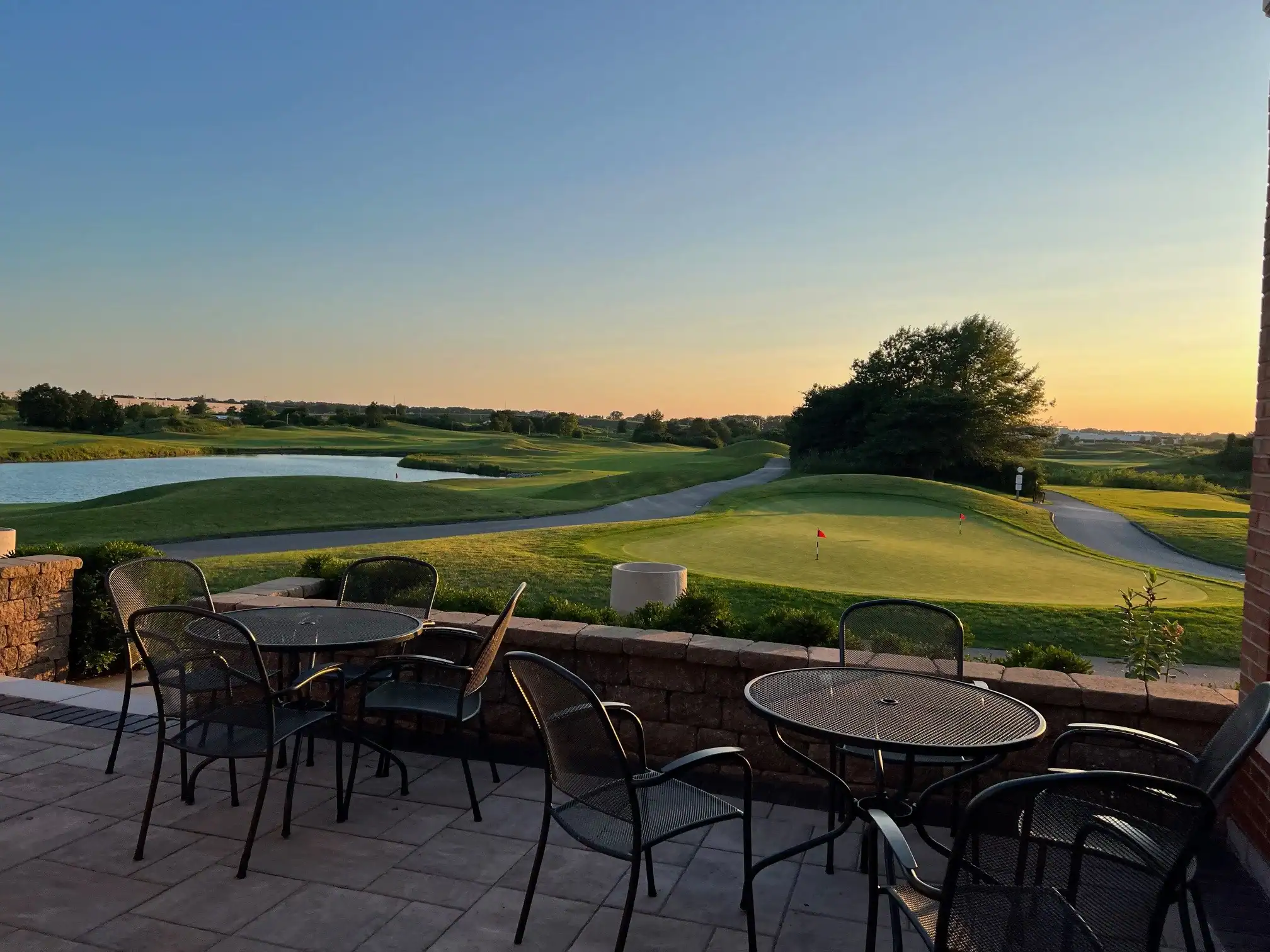 A golf course at sunset, viewed from a patio with outdoor tables and chairs. The sky is blue and orange, and a green putting area with a red flag is visible.