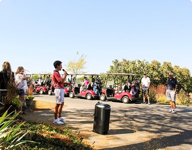 A young man in a red shirt speaks into a microphone in front of a line of red golf carts filled with golfers, under a clear blue sky.
