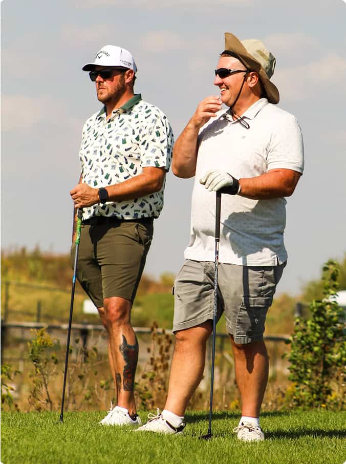 Two men in golf attire stand on a green golf course under a sunny sky, holding golf clubs. The man on the left wears a white cap and has a leg tattoo. The man on the right wears a bucket hat and is smiling.