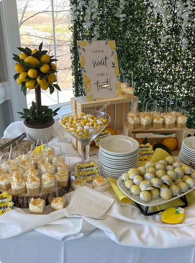 A vibrant lemon-themed dessert table featuring a lemon topiary, various individual desserts in cups, yellow and white candies, lemon-shaped cookies, and white chocolate truffles, all set against a green foliage wall.