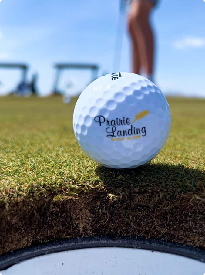 A white golf ball with the 'Prairie Landing Golf Club' logo balanced on the very edge of a golf hole, with a golfer and golf carts blurred in the sunny background.
