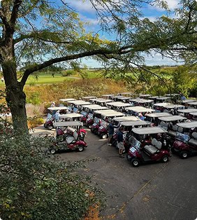 An elevated view of dozens of red golf carts parked in rows on a paved lot at a golf course, with people gathered around them and trees framing the scene.