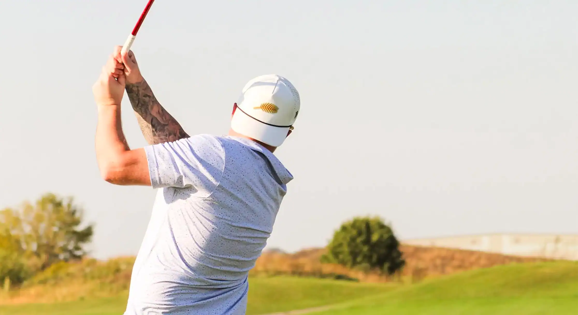 A golfer, seen from behind, in the follow-through of a golf swing. They wear a white hat with a gold leaf emblem and a speckled light-colored shirt, holding a red-shafted club against a bright sky and green golf course.