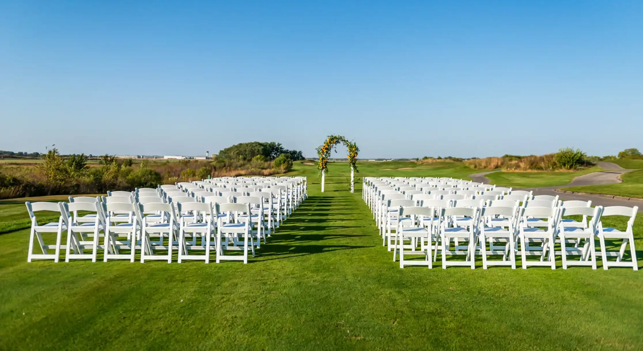 An outdoor wedding ceremony setup on a green lawn. White chairs are arranged in two sections facing a floral arch adorned with yellow and orange flowers, under a clear blue sky.