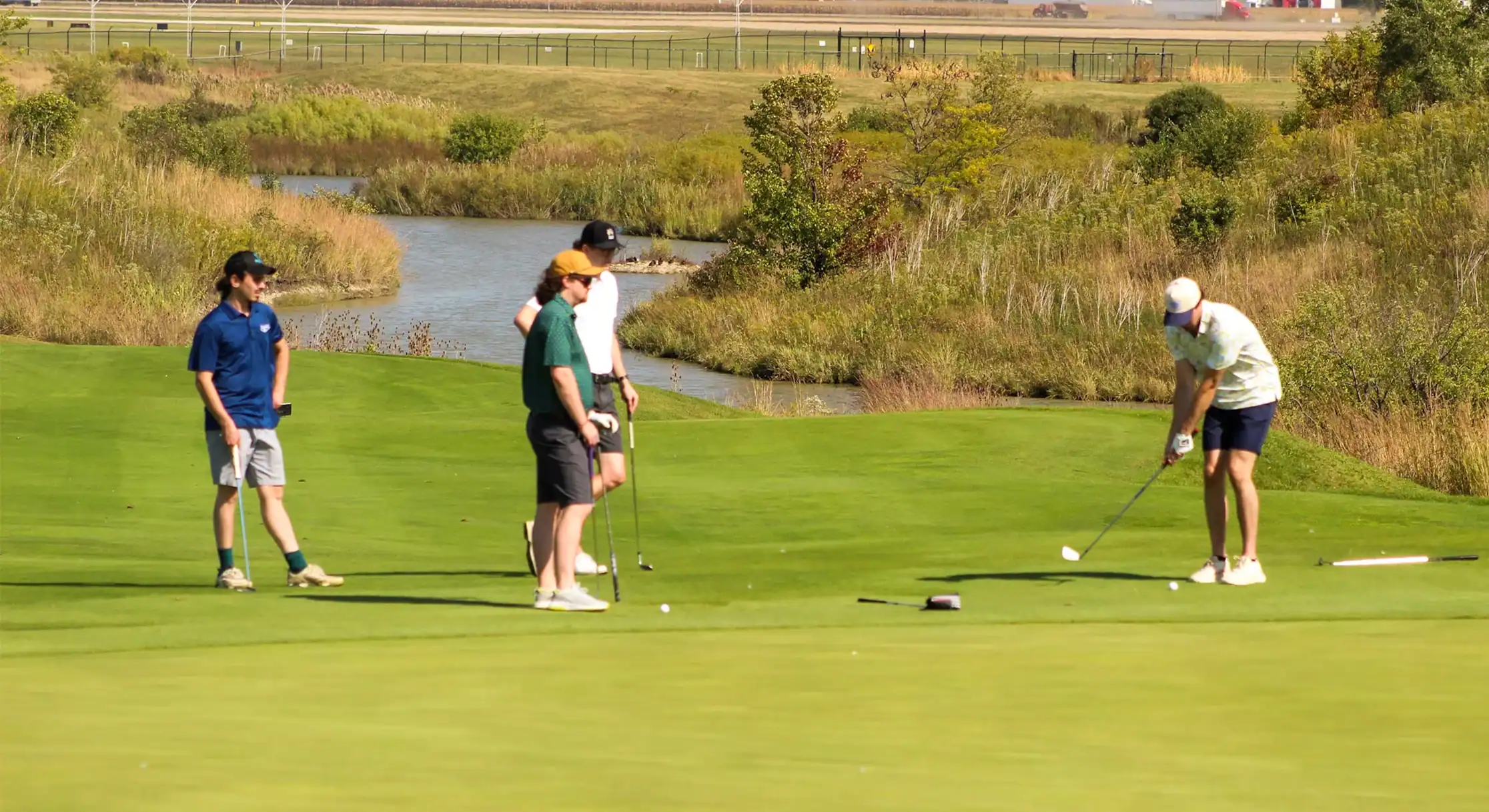 Four men golfing on a sunny day. One man in a patterned shirt is about to hit the ball, while three others watch on a green course with a pond and trees in the background.