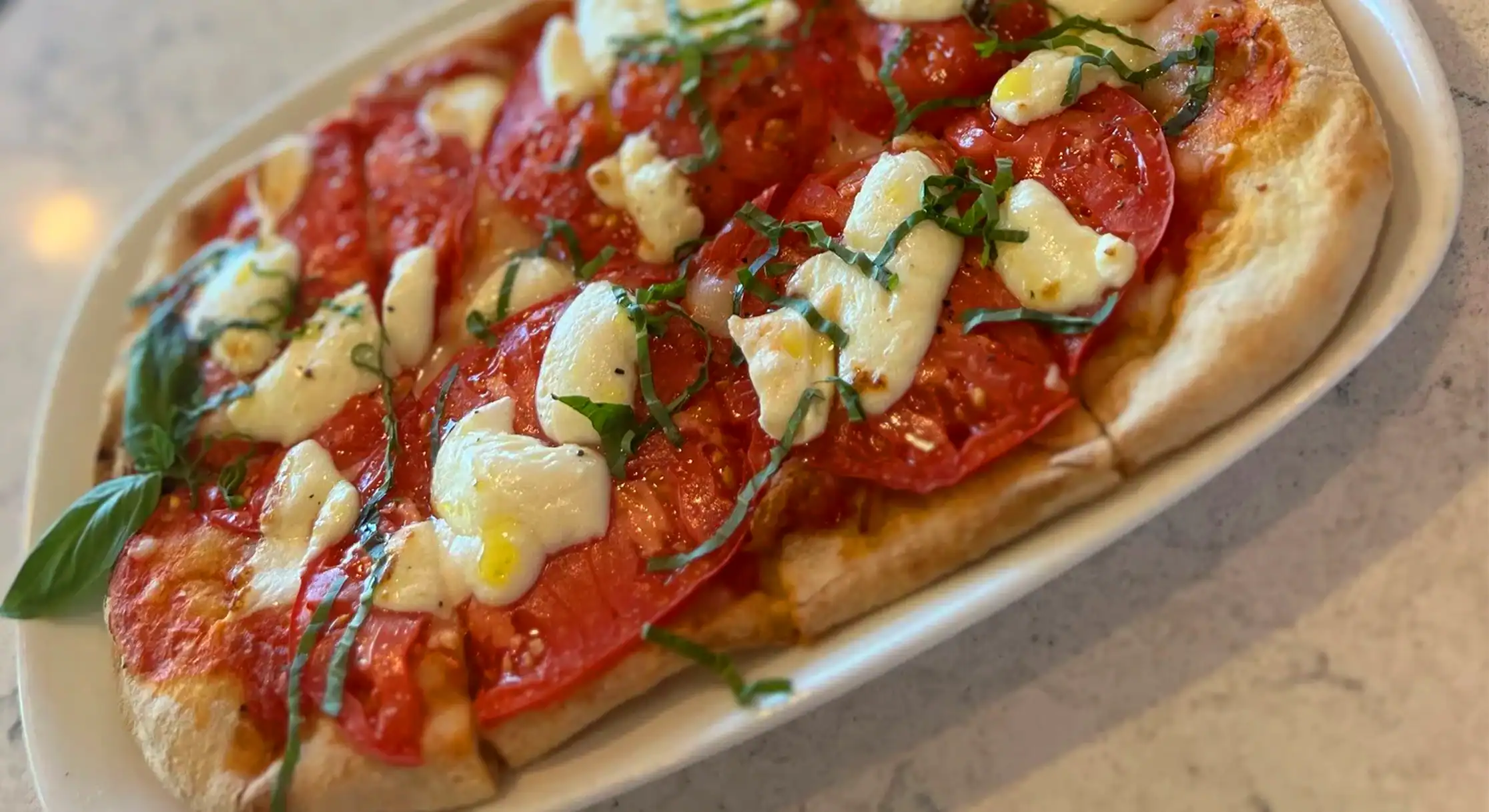 A rectangular flatbread pizza topped with large slices of red tomatoes, dollops of white mozzarella cheese, and fresh green basil, served on a white platter.