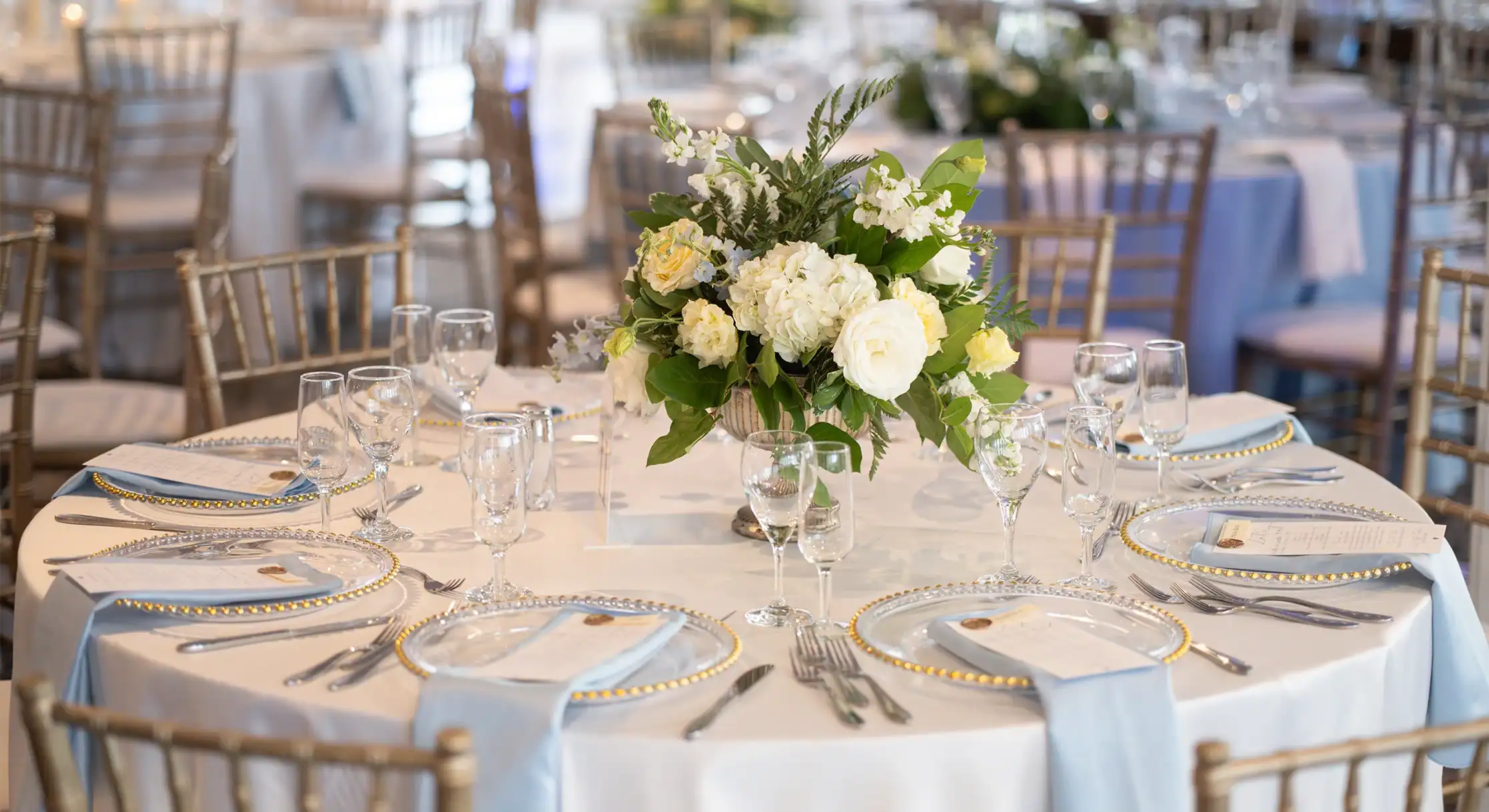 A round table set for a formal event with a white tablecloth, light blue napkins, gold-rimmed plates, and a centerpiece of white and yellow flowers. Gold Chiavari chairs are visible around the table and in the blurred background.