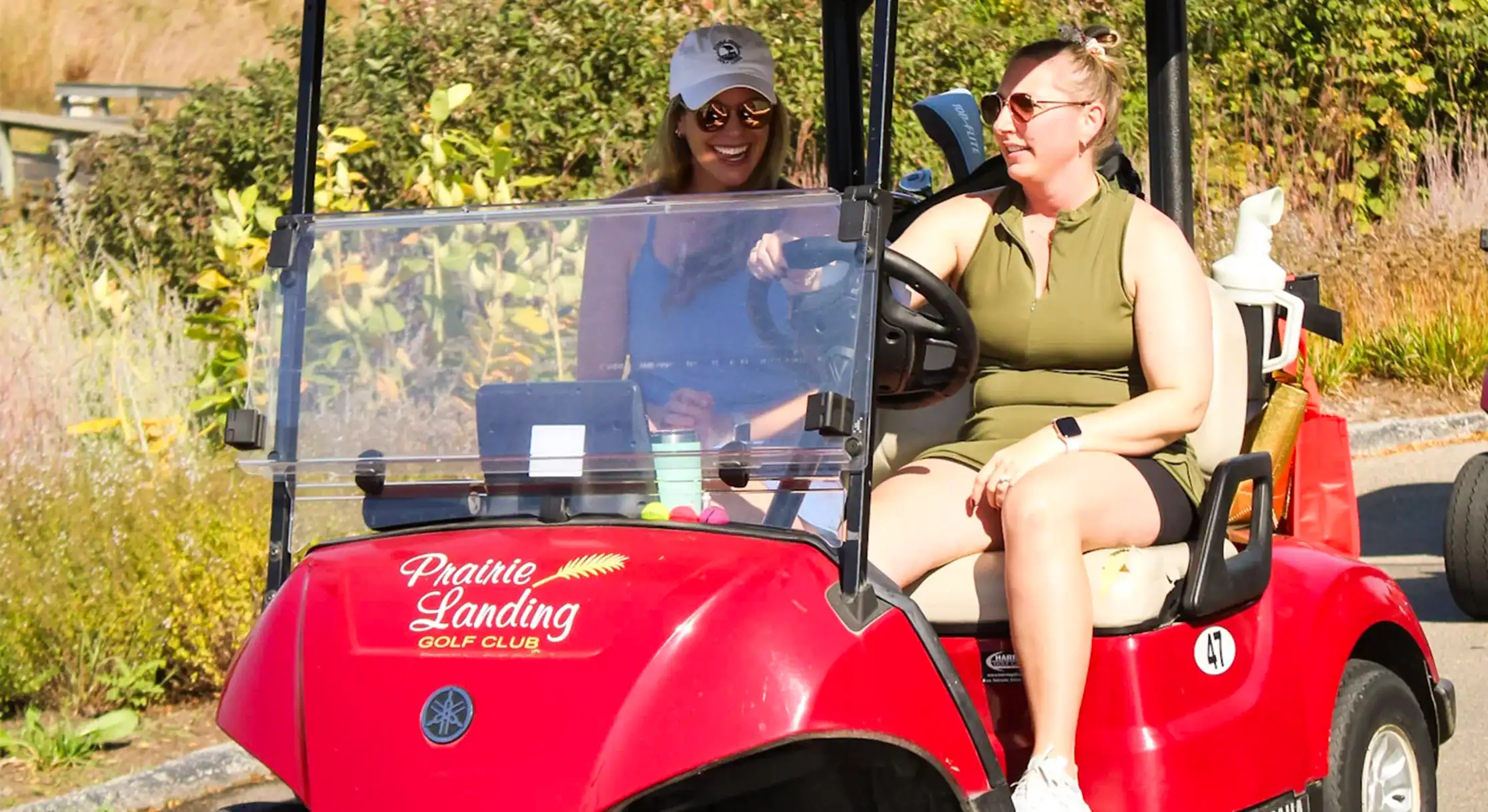 Two smiling women in a red golf cart on a sunny day. One drives in an olive top, the other, in a blue top and white cap, is the passenger.