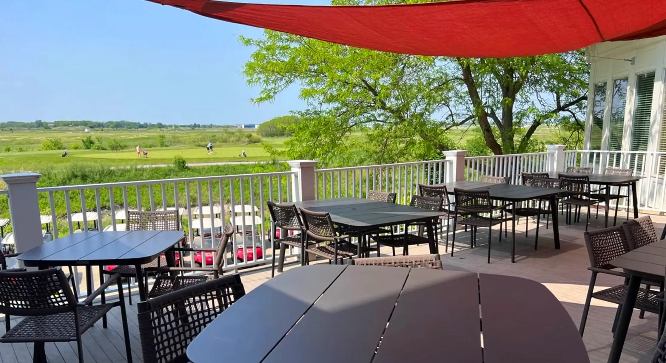 An outdoor patio with dark dining tables and chairs under a red sunshade, overlooking a green golf course with golfers and golf carts on a sunny day.