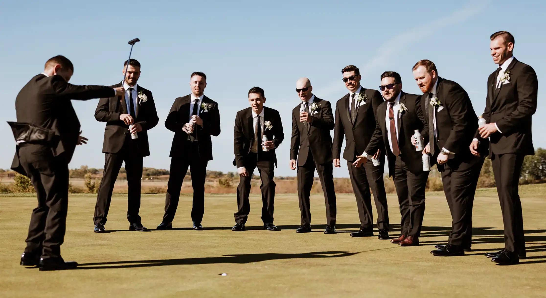 Nine groomsmen in black suits on a golf putting green. One man is putting, while the others watch, holding drinks and smiling under a clear blue sky.