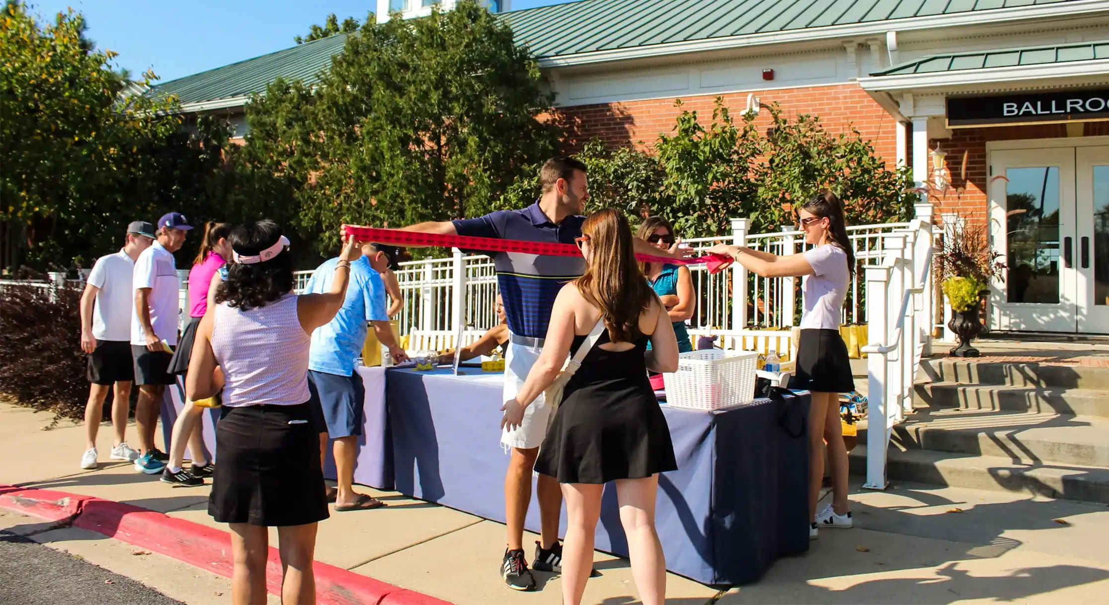 A group of people at an outdoor event, likely golf registration, with a man and two women holding a long red ribbon. A building with a 'BALLROOM' sign is in the background.
