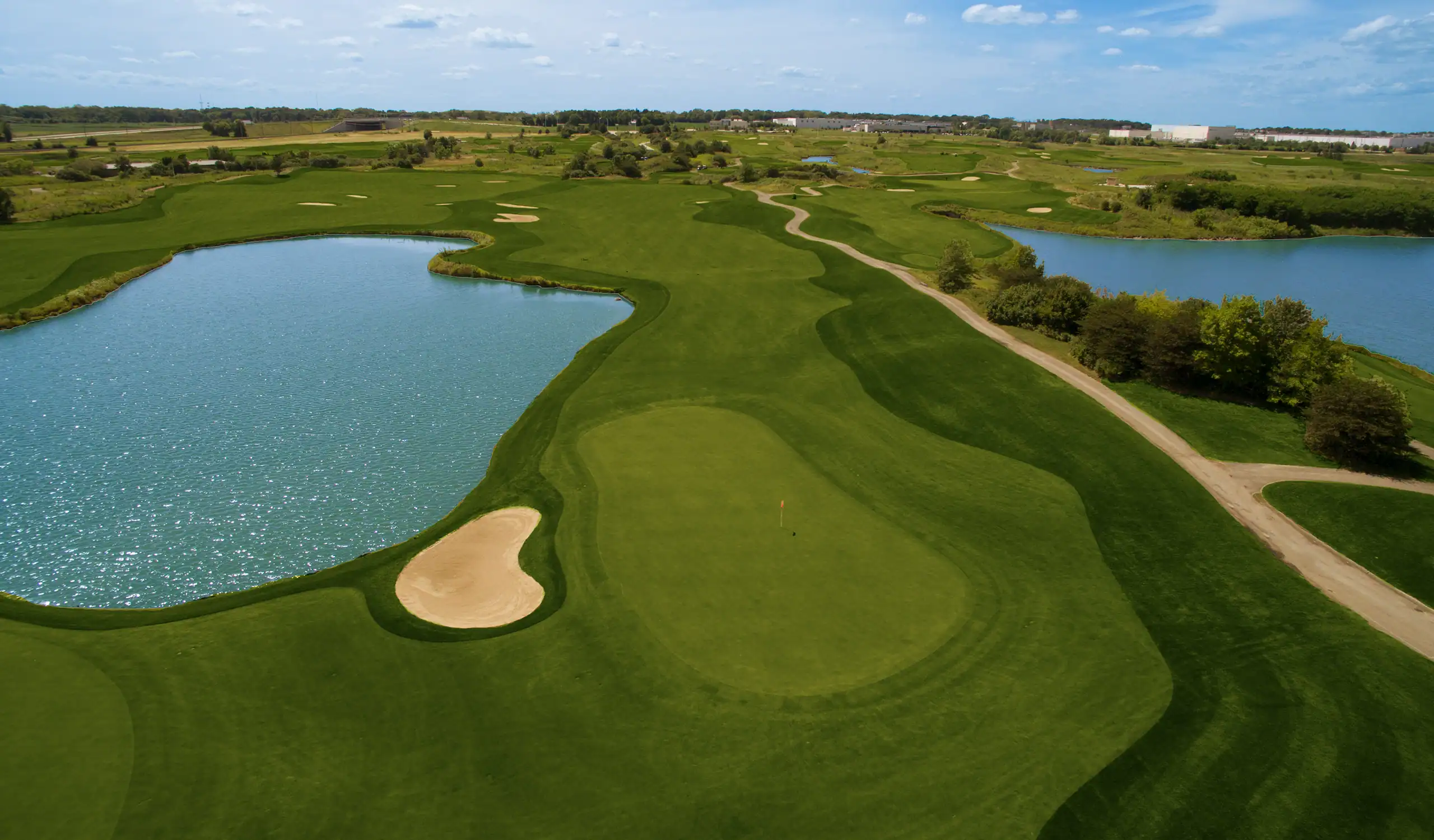 Aerial view of a lush green golf course with two large blue water hazards, a sand bunker, and a putting green with a red flagstick, under a partly cloudy sky.